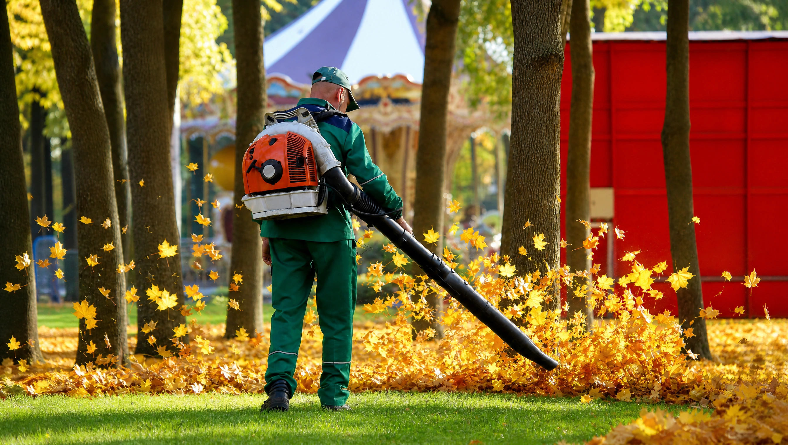Landscaper in green uniform using a backpack leaf blower to clear fallen autumn leaves from a maintained lawn